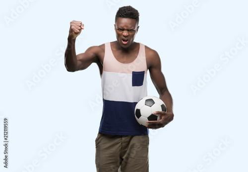 Young african american man holding soccer football ball annoyed and frustrated shouting with anger, crazy and yelling with raised hand, anger concept © Krakenimages.com