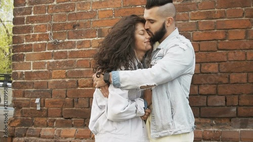 Beautiful romantic young couple outdoor with the young man hugging his girlfriend or wife and kisses her with great love wearing denim jackets near brick wall, slow motion