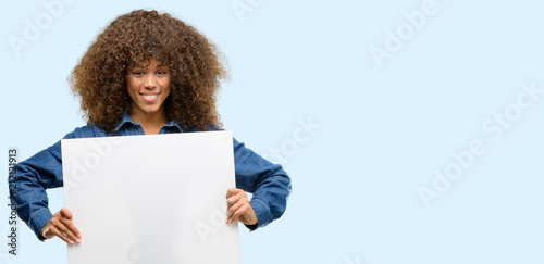 African american woman wearing blue jumpsuit holding blank advertising banner, good poster for ad, offer or announcement, big paper billboard photo
