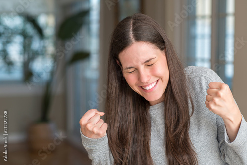 Young beautiful woman at home very happy and excited doing winner gesture with arms raised, smiling and screaming for success. Celebration concept.