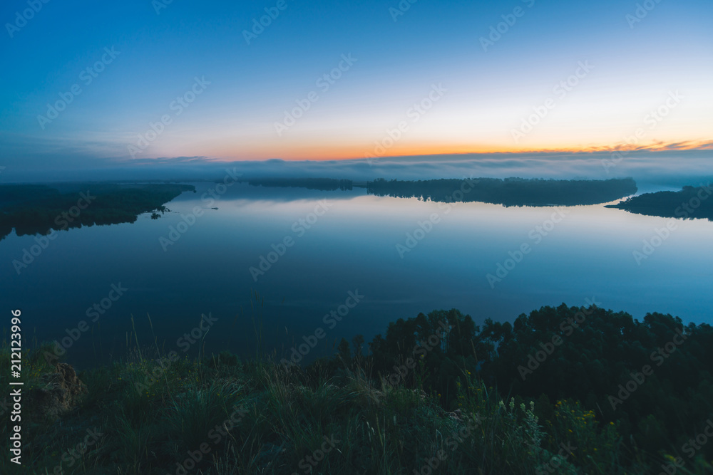 Mystical view from high shore on river. Riverbank with forest under mist. Early haze above trees. Orange glow in picturesque predawn sky. Colorful morning atmospheric calm landscape of majestic nature