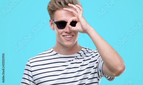 Young handsome blond man wearing sunglasess with happy face smiling doing ok sign with hand on eye looking through fingers
