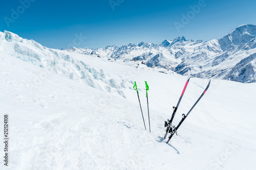A pair of skis and ski poles stick out in the snow on the mountain slope of the Caucasus against the backdrop of the Caucasian mountain range and the blue sky on a sunny day