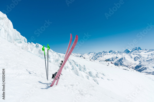 A pair of skis and ski poles stick out in the snow on the mountain slope of the Caucasus against the backdrop of the Caucasian mountain range and the blue sky on a sunny day
