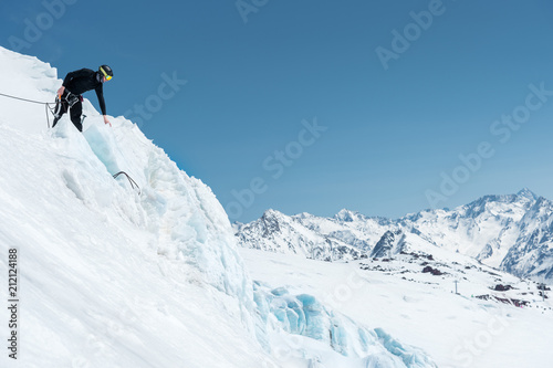 A professional mountaineer in a helmet and ski mask on insurance makes a nick-hole in the glacier against the backdrop of the Caucasian snow-capped mountains © yanik88