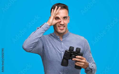 Handsome young man looking through binoculars with happy face smiling doing ok sign with hand on eye looking through fingers