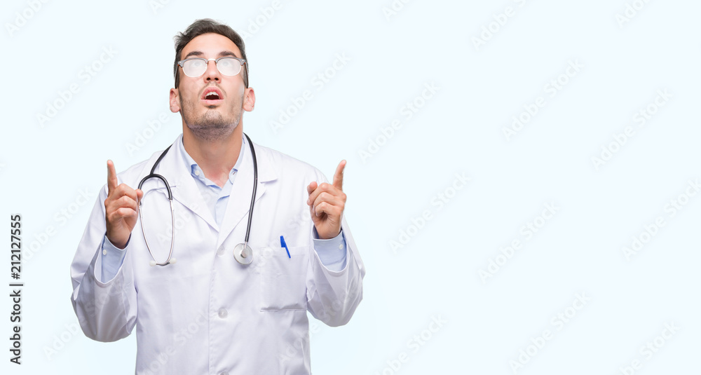 Handsome young doctor man amazed and surprised looking up and pointing with fingers and raised arms. Handsome young doctor man amazed and surprised looking up and pointing with fingers and raised arms.