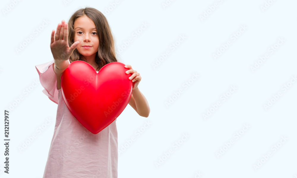 Brunette hispanic girl holding red heart with open hand doing stop sign with serious and confident expression, defense gesture Brunette hispanic girl holding red heart with open hand doing stop sign with serious and confident expression, defense gesture