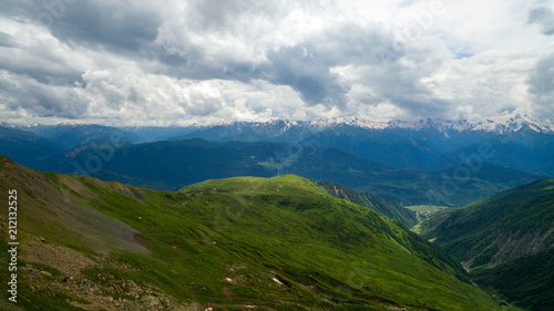 green slope against the background of snowy peaks of mountains and cloudy sky © Aleksei Lazukov