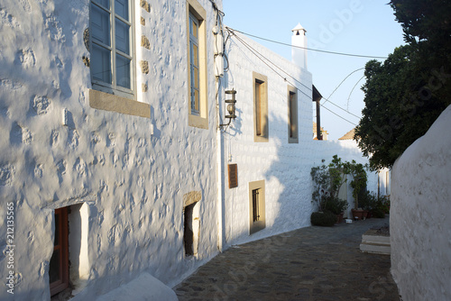 Fototapeta Naklejka Na Ścianę i Meble -  A view of a narrow street with arch and wooden windows and doors with white wall stone architecture of the island Patmos, Greece