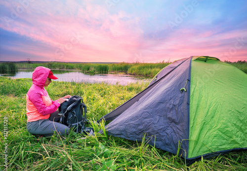 Girl in pink jacket opens her backpack near tent on the river ba