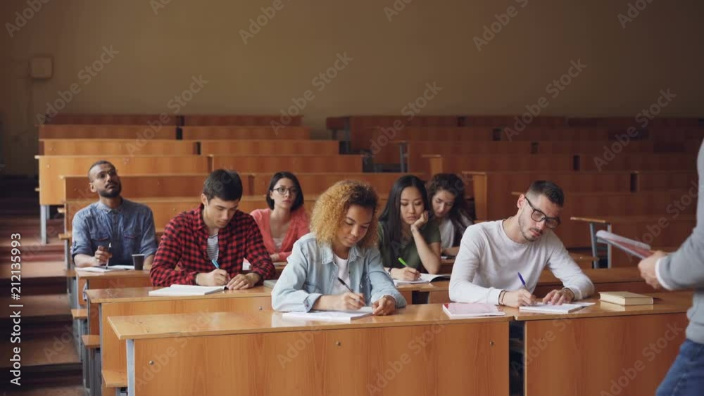 Professor is reading lecture to group of multiracial students standing in classroom with papers and speaking while young people are listening to him and writing.
