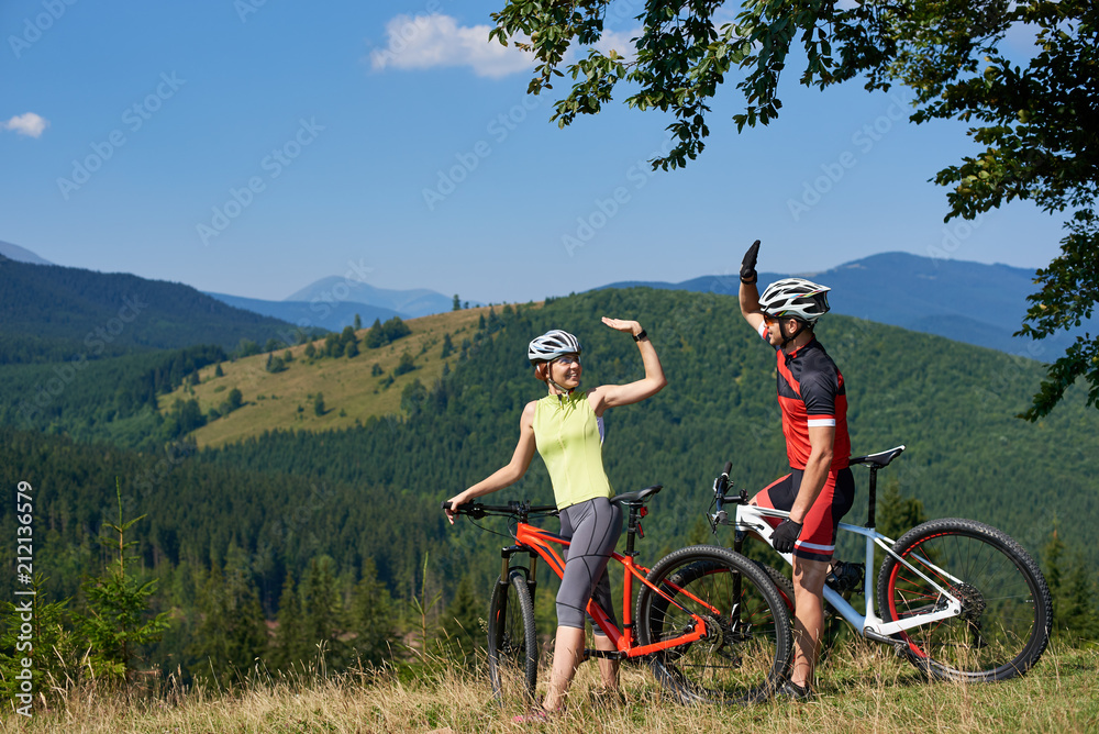 Obraz premium Young smiling bikers couple, man and woman, standing with bikes on grassy hill under big tree having fun with raised hands on fantastic magnificent mountain covered with forest summer background.