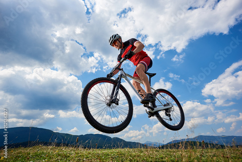 Athletic sportsman cyclist in professional sportswear and helmet flying in air on his bicycle on summer blue sky with white clouds and distant mountains on background. Outdoor extreme sport concept