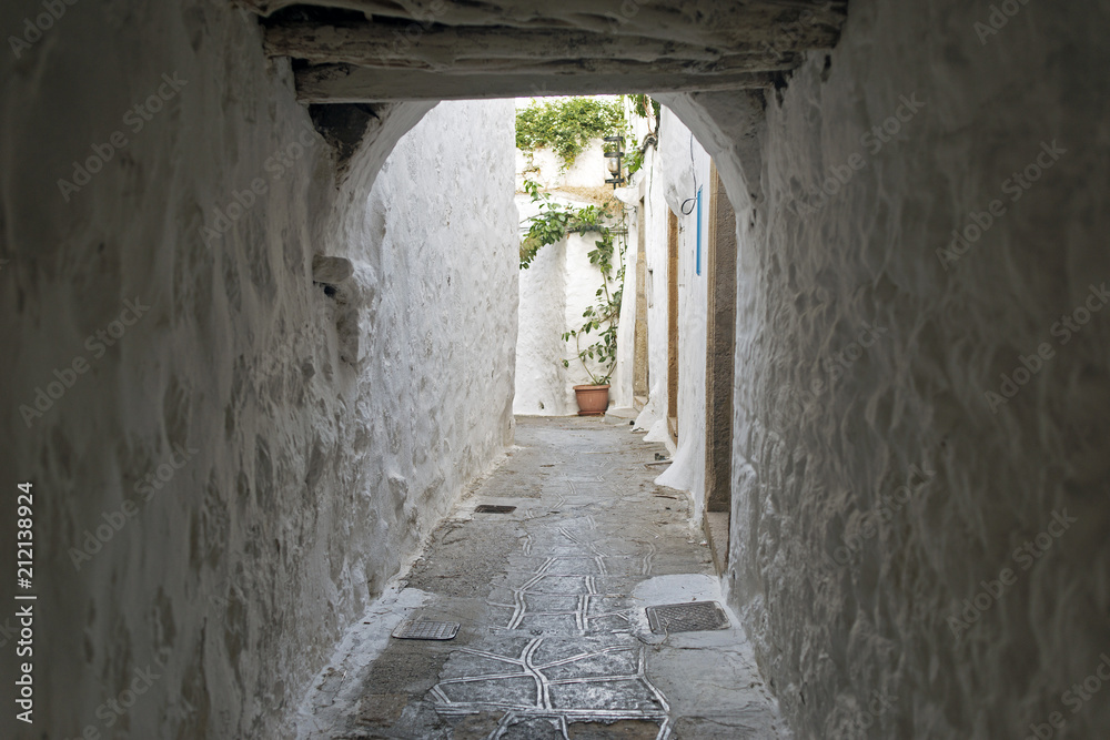 Fototapeta premium A view of a narrow street with arch and wooden windows and doors with white wall stone architecture of the island Patmos, Greece