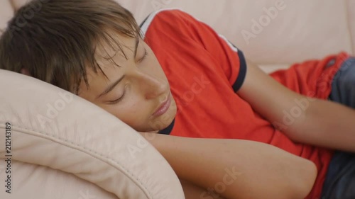Teenager sleeping on couch. Close-up of caucasian teen boy in red t-shirt and blue jeans sleeping on beige leather sofa in daytime. Lack of sleep