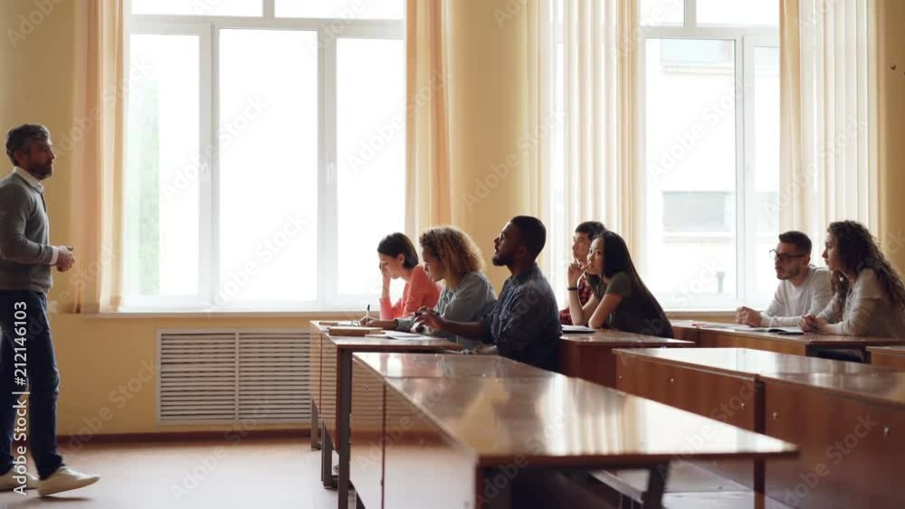 Friendly teacher is talking to students, smart mixed race man is raising hand and speaking sitting at table in lecture hall while his classmates are writing. Friendly teacher is talking to students, smart mixed race man is raising hand and speaking sitting at table in lecture hall while his classmates are writing.