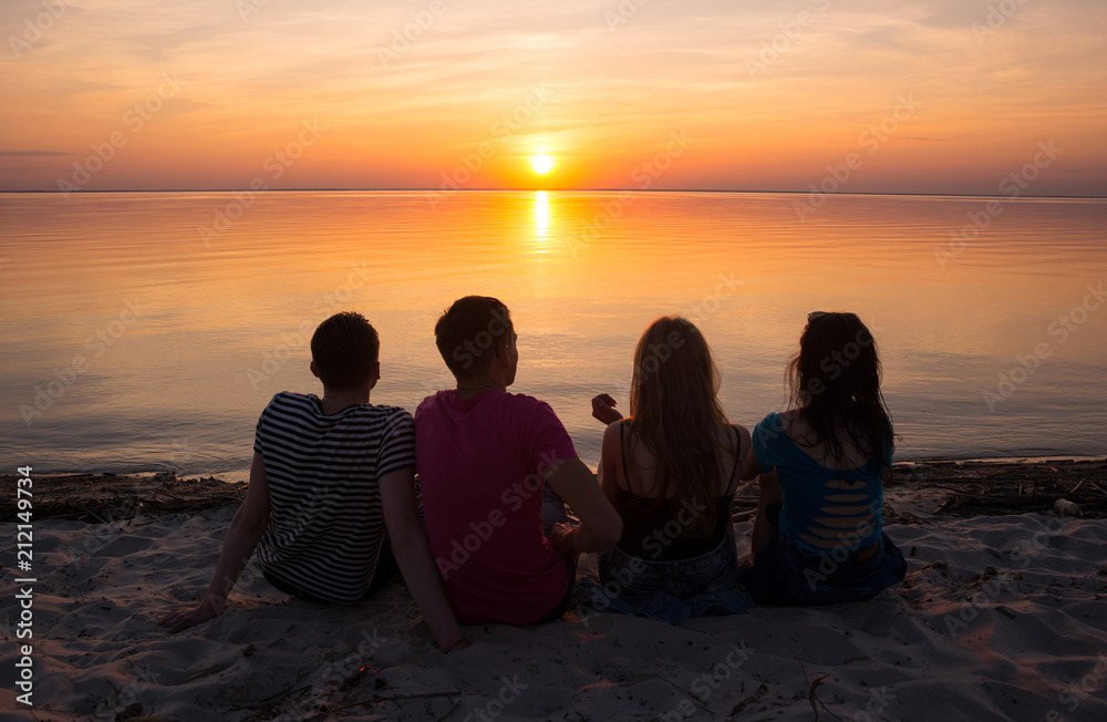 Young people - guys and girls - sit on the beach and watch the sunset.