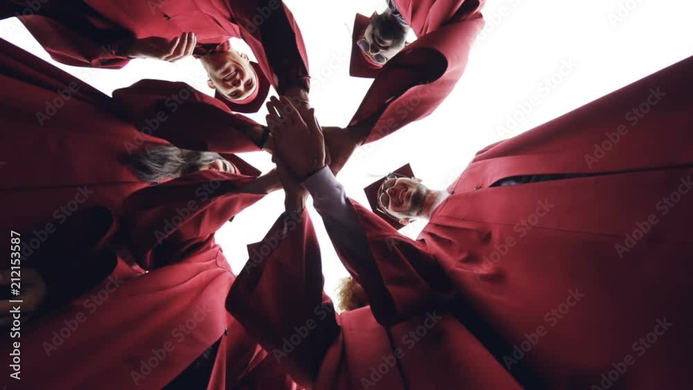 Low angle shot of graduating students putting hands together then raising them in the air and clapping hands, young people are wearing traditional gowns and hats.