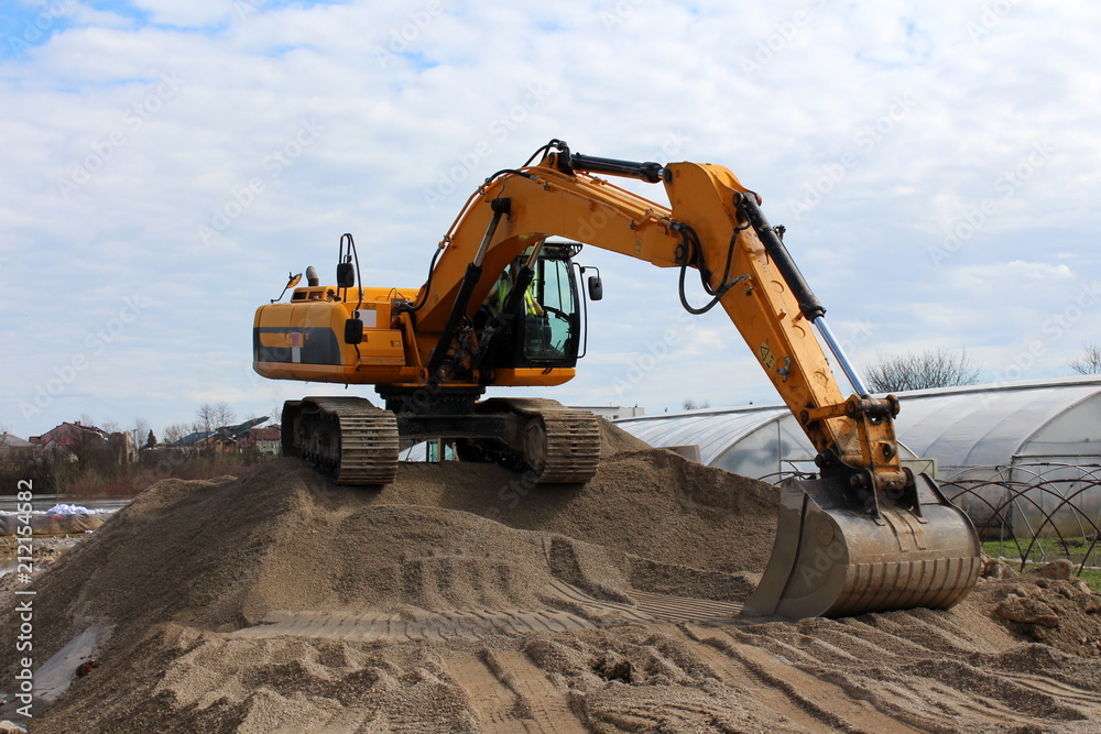 Excavator standing on top of sand pile waiting for truck with greenhouse, houses, river and cloudy blue sky in background Excavator standing on top of sand pile waiting for truck with greenhouse, houses, river and cloudy blue sky in background
