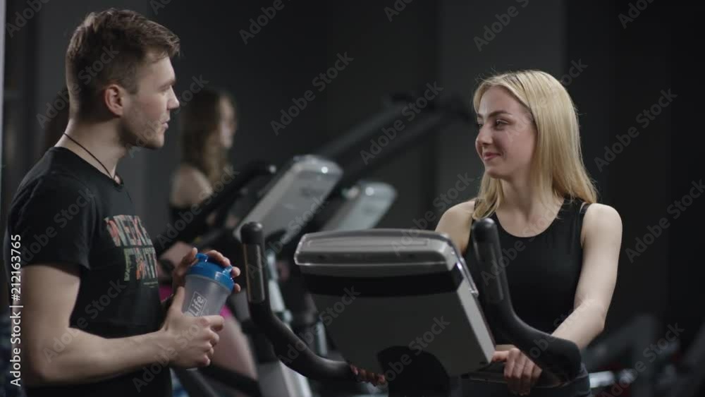 nice girl in blak sport wear vigorously works on exercise bike and guy comes to meet and to ask her name in the new gym against the background of engaged girls nice girl in blak sport wear vigorously works on exercise bike and guy comes to meet and to ask her name in the new gym against the background of engaged girls