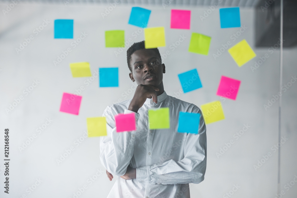 Young afro american business man standing in front of stickers glass wall and looking on futures plans at his office place Young afro american business man standing in front of stickers glass wall and looking on futures plans at his office place