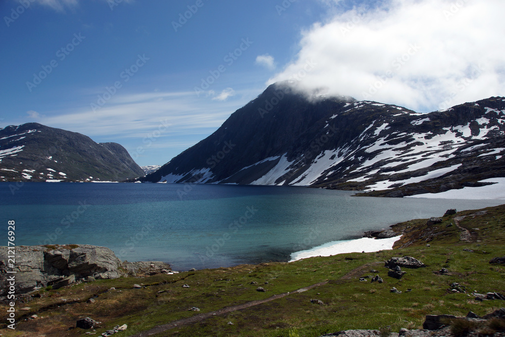 The blue lake lies at the foot of the mountains covered with snow in the summer in Norway