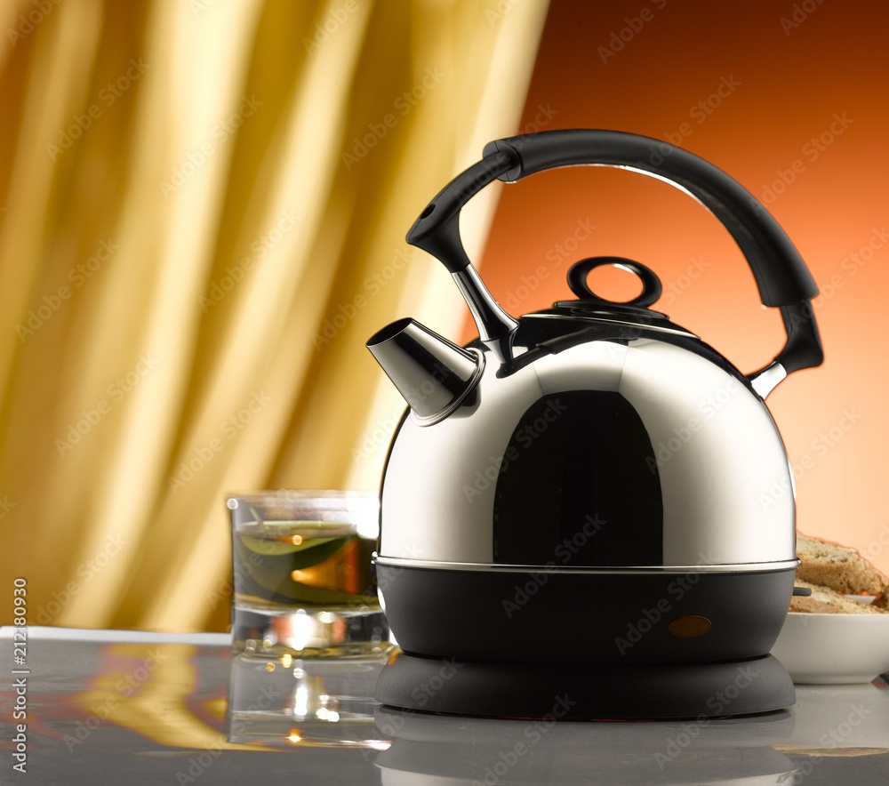 Electric kettle and tea glass on a table. Yellow curtain and brown wall in  the background Stock Photo