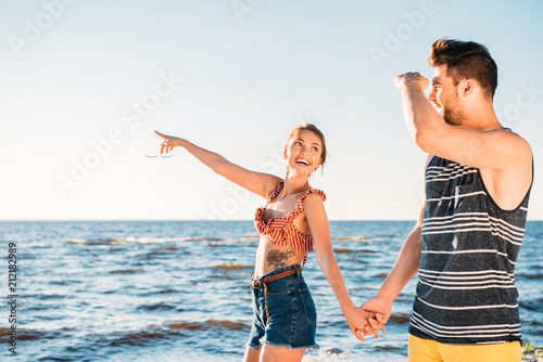 happy young couple holding hands and looking away while walking together on beach