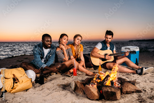 young multiethnic friends enjoying guitar and spending time together on sandy beach at sunset