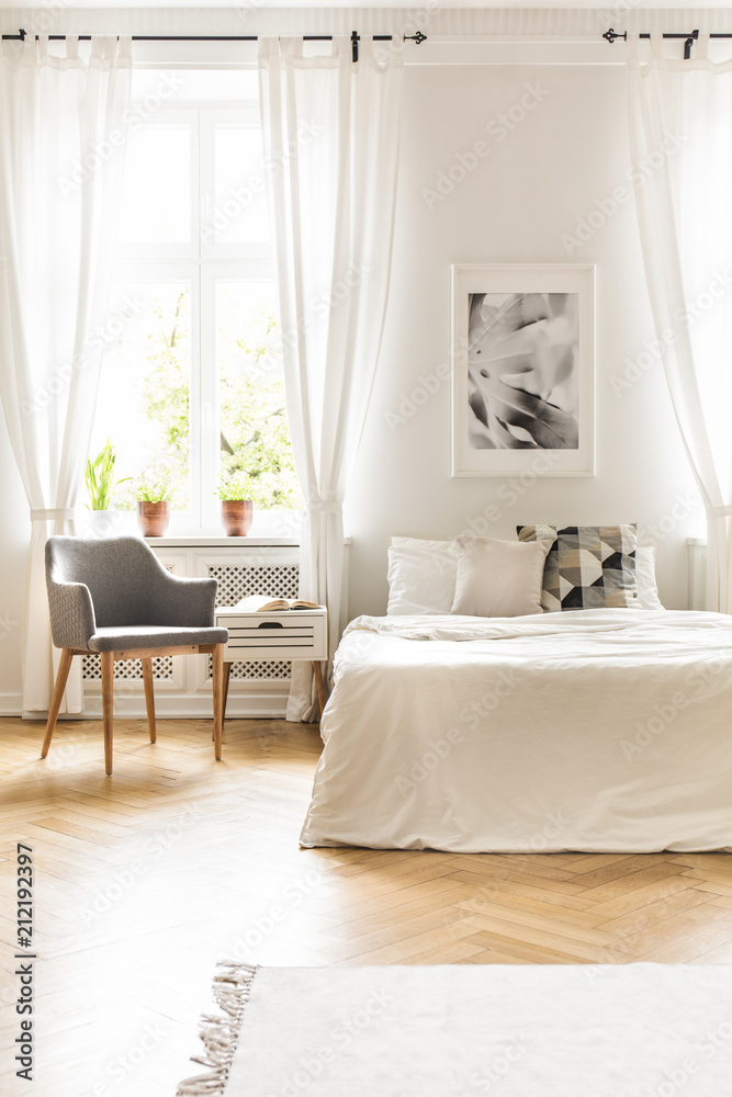 Grey armchair at window with drapes in white bedroom interior with poster above bed. Real photo