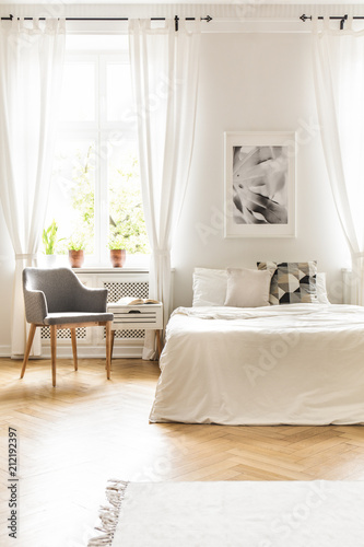 Grey armchair at window with drapes in white bedroom interior with poster above bed. Real photo