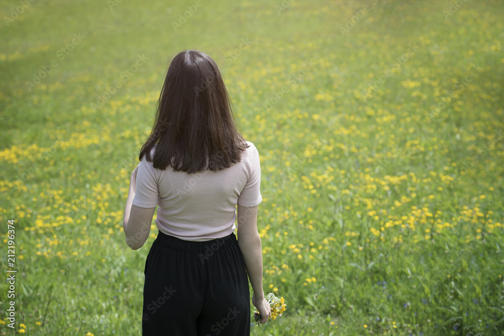 Young woman is standing on a flower meadow. Summer sunny day. Back view Young woman is standing on a flower meadow. Summer sunny day. Back view