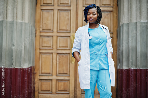 Stylish african american doctor with stethoscope and lab coat posed against door of hospital.