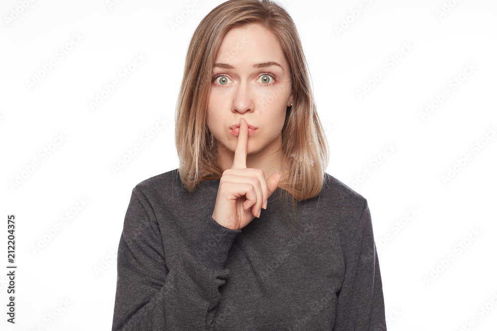 Shot of beautiful female with long blonde dyed hair wears casual long-sleeved grey t-shirt, shows hush sign, has serious expression, asks not to tell her secret anybody, hopes for loyalty and silence. Shot of beautiful female with long blonde dyed hair wears casual long-sleeved grey t-shirt, shows hush sign, has serious expression, asks not to tell her secret anybody, hopes for loyalty and silence.