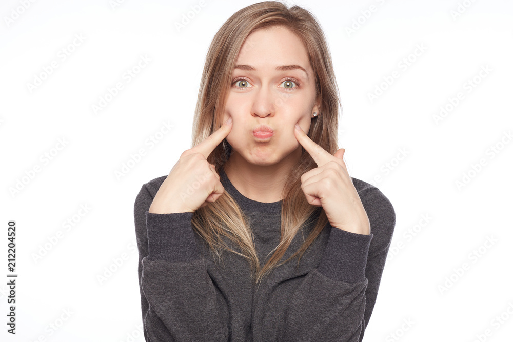 Indoor shot of Caucasian pretty blond woman making grimace blowing cheeks, having pleased face expression. Beautiful young woman with clean tender skin dressed in casual clothes mocking at camera. Indoor shot of Caucasian pretty blond woman making grimace blowing cheeks, having pleased face expression. Beautiful young woman with clean tender skin dressed in casual clothes mocking at camera.