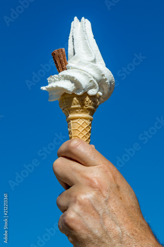 A hand holding a vanilla ice cream cone against a clear blue sky