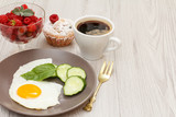 Plate with fried egg, fresh cut cucumber and leaf of basil, fork, cup of black coffee, muffin and glass bowl with fresh raspberries