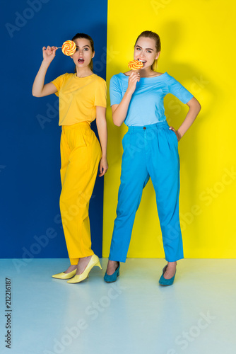 Elegant stylish women eating lollipops on blue and yellow background