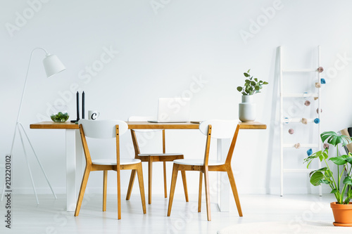 Bright and natural start-up office interior with a laptop and three wooden chairs around a simple desk. Real photo