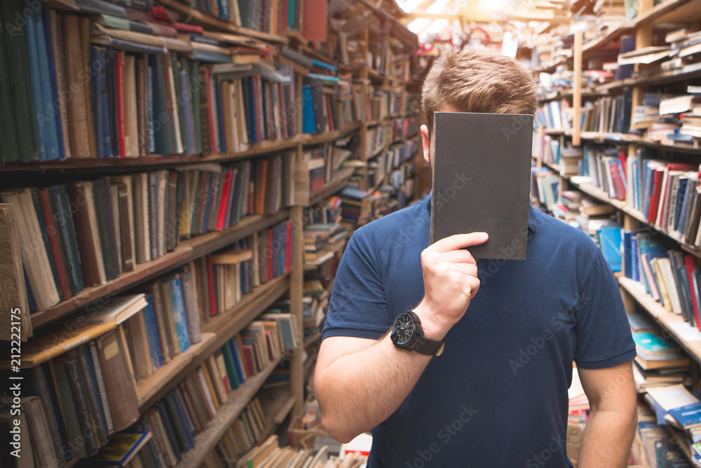 Portrait of a man standing in an atmospheric public library and closing his face with a book. A student in a library with a book on the face. Facebook Portrait of a man standing in an atmospheric public library and closing his face with a book. A student in a library with a book on the face. Facebook