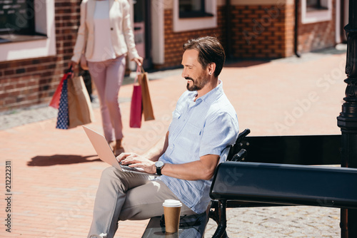 handsome man sitting on bench with laptop, woman walking with shopping bags on street