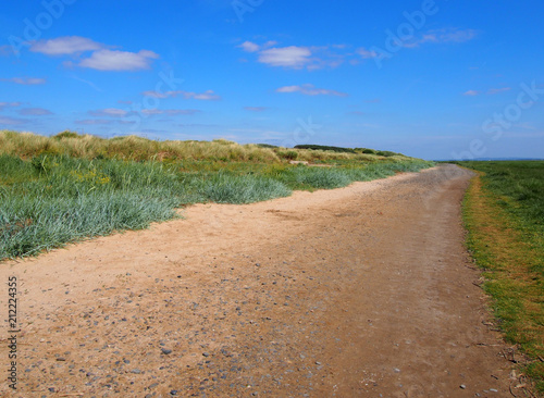 single land dirt road mad of sand and stones extending to the distance with grass covered dunes and bright blue summer sky near the ribble estuary in lancashire england