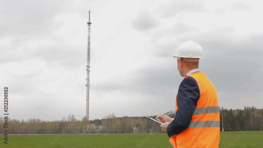 A man in a signal jacket and helmet writes notes in the tablet, in the background a telephone mobile tower