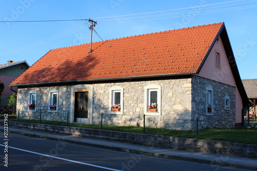 Renovated small stone house with new red roof at sunset with tree shadows and grass in front next to local road