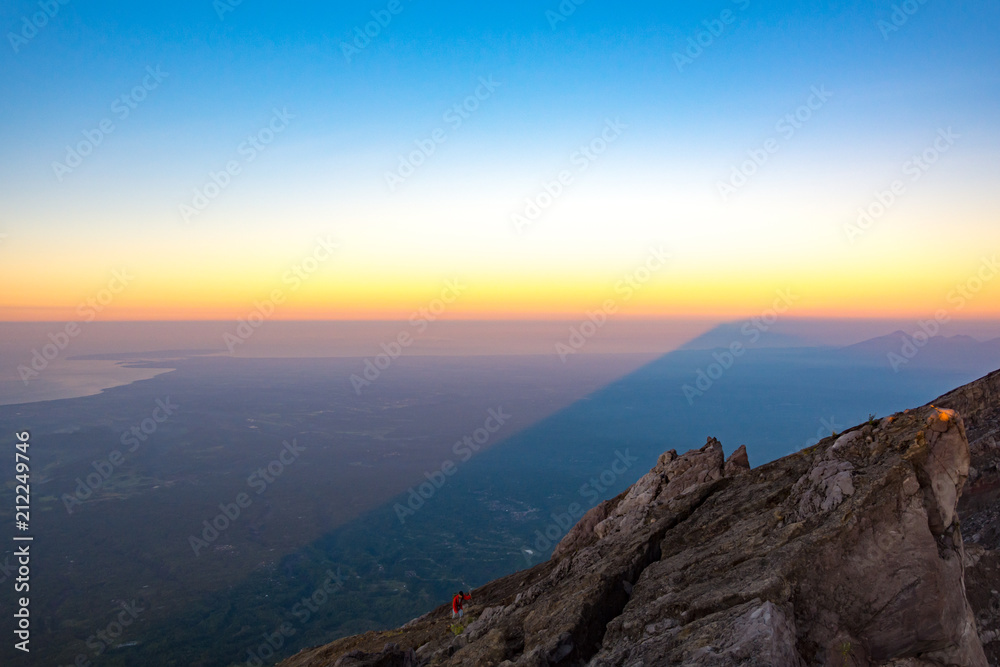 A Man In Red Jacket is Climbing to The Top of Mount Agung Volcano, The Active Volcano in Bali, Indonesia at Sunrise with A Shadow of Mount Agung Over Bali Background.