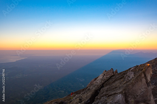 A Man In Red Jacket is Climbing to The Top of Mount Agung Volcano  The Active Volcano in Bali  Indonesia at Sunrise with A Shadow of Mount Agung Over Bali Background.