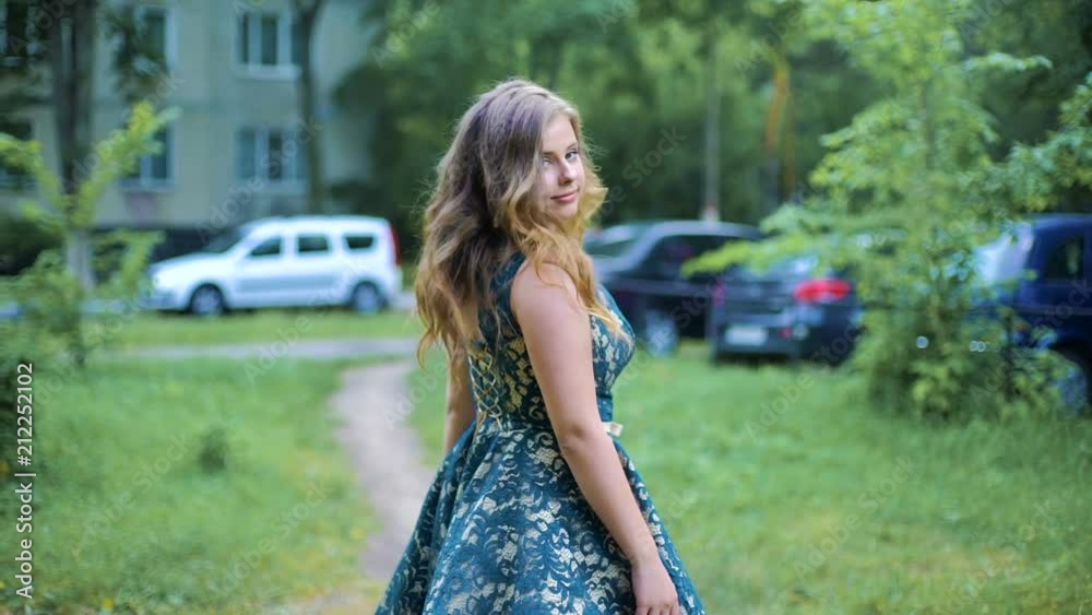 Back view of young woman walking through the street near the tree. Hair waving on wind. The girl turns around. Slow mo.