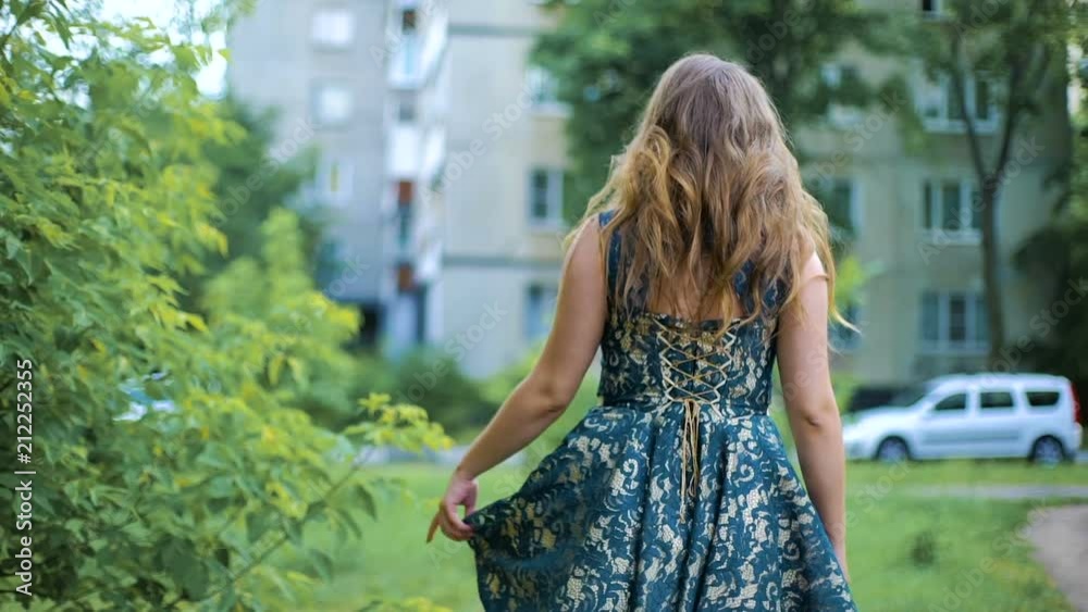 Back view of young woman walking through the street near the tree. Hair waving on wind. The girl turns around. Slow mo. Back view of young woman walking through the street near the tree. Hair waving on wind. The girl turns around. Slow mo.