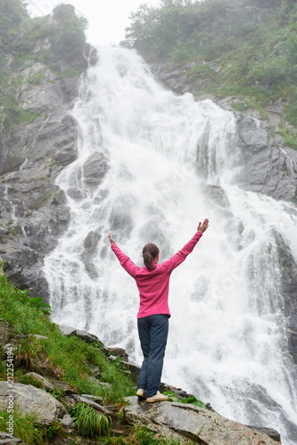 Pretty young girl standing with raised hands at the big powerful waterfall. Beautiful woman enjoying nature in warm autumn weather. Traveling to the waterfall Balea in Romanian Fagarash mountains.